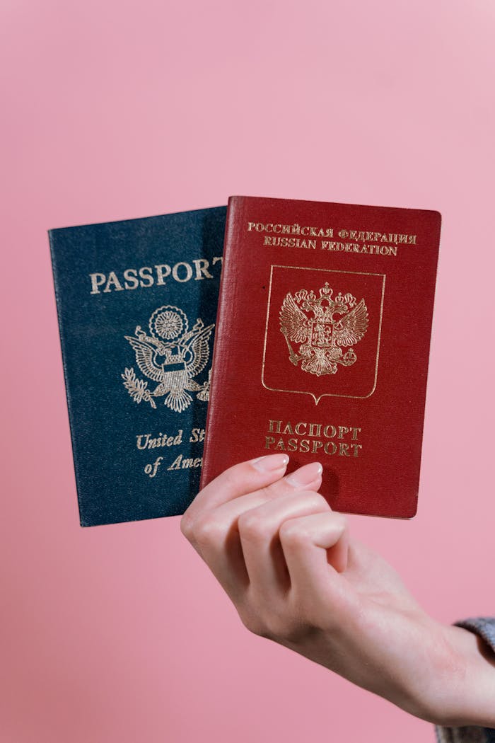 A hand holding US and Russian passports against a pink background.